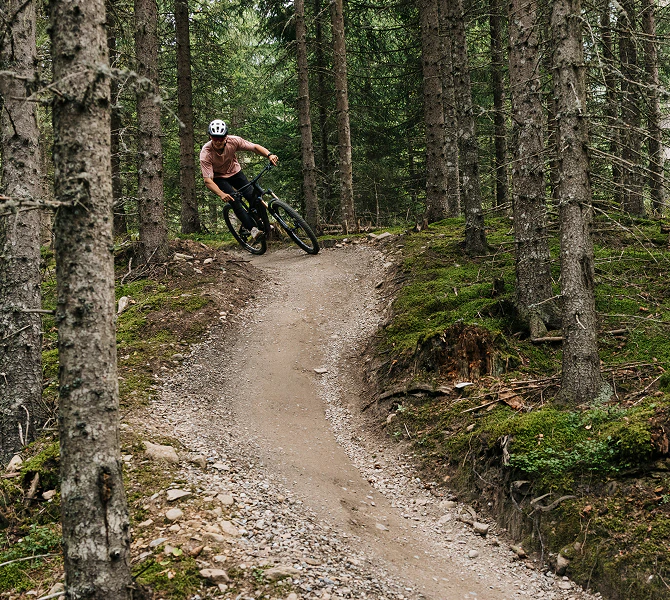 Mountainbiker fährt eine steile Kurve auf einem Waldtrail, umgeben von Nadelbäumen.