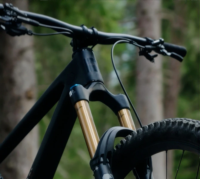 Nahaufnahme des matt-schwarzen Rokua 170 Ultra Mountainbike-Cockpits mit goldener Fox-Federgabel vor einem unscharfen Waldhintergrund.