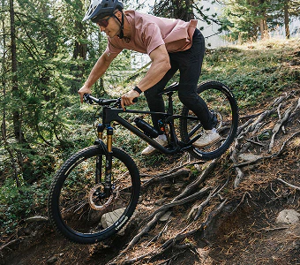 Mountainbiker fährt mit einem schwarzen Raymon Rokua Bike über einen wurzeligen Waldpfad.