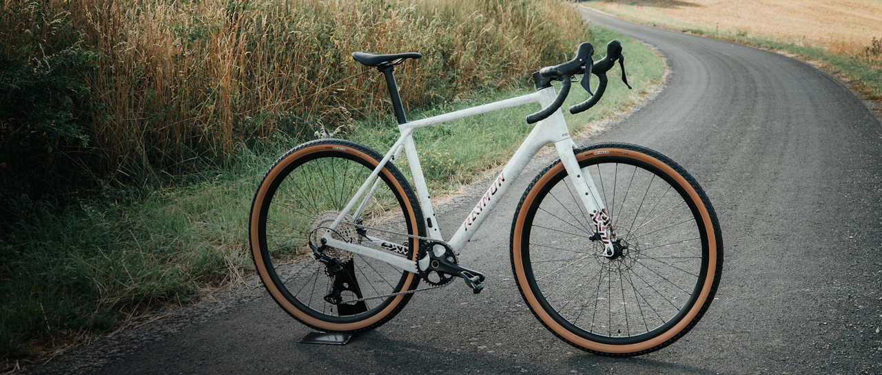 Weißes RAYMON SORENO Comp Gravel-Bike mit braunen Reifenflanken steht auf einer asphaltierten Landstraße zwischen Feldern.