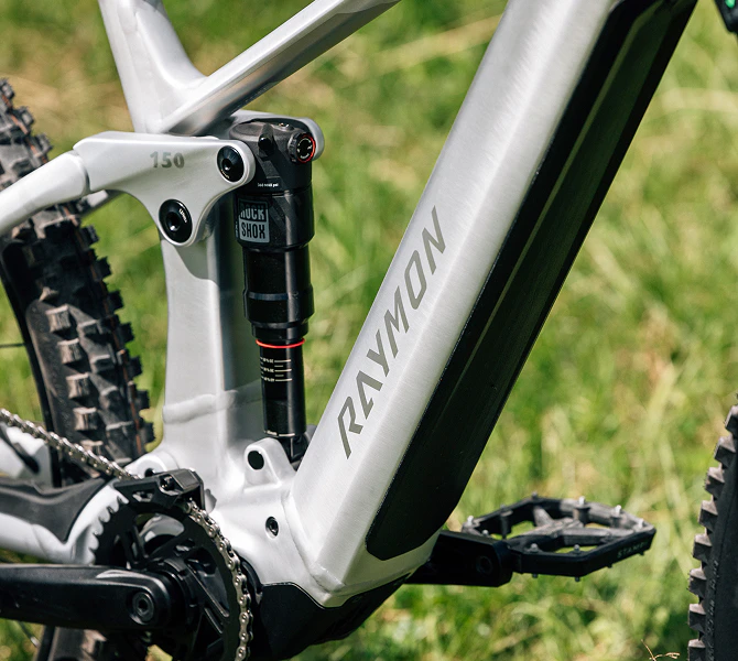 Nahaufnahme des Rahmens eines silbernen Raymon E-Bikes mit RockShox Dämpfer auf einer grünen Wiese.
