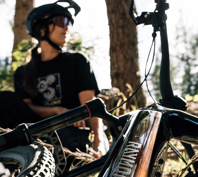 Nahaufnahme eines Raymon E-Bikes im Wald mit einer Mountainbikerin im Hintergrund.