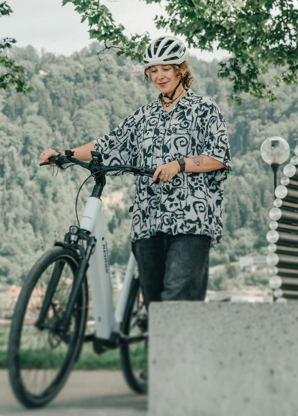 Frau mit Fahrradhelm schiebt ein weißes Raymon Numa E-Bike in einer bewaldeten Umgebung.
