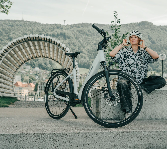 Frau setzt sich neben einem weißen Raymon Numa E-Bike auf eine Granitbank und setzt ihren Fahrradhelm ab.