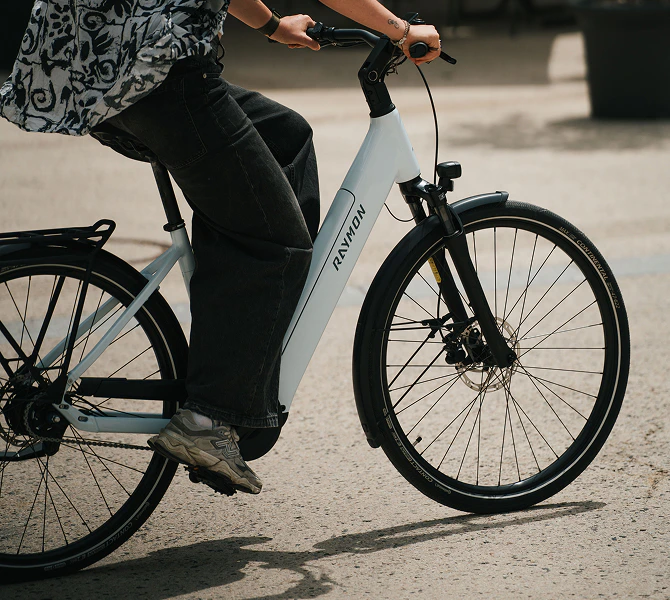Person in schwarzer, weiter Jeans fährt auf einem weißen Raymon Numa E-Bike über einen asphaltierten Platz.