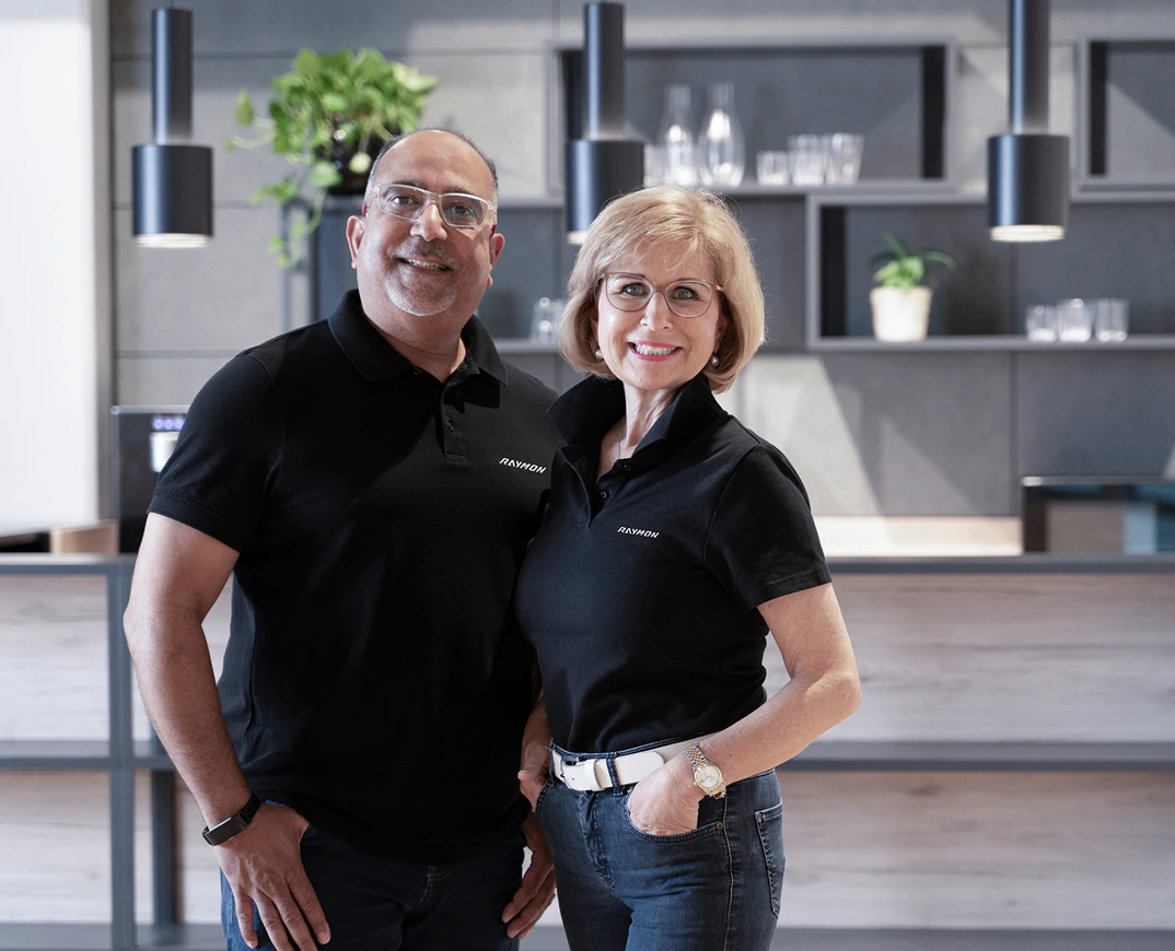 Susanne und Felix Puello lächeln in schwarzen Raymon Poloshirts vor einer modernen Bürokulisse.