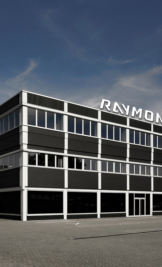 Modernes Bürogebäude der Firma Raymon mit großem Logo auf dem Dach unter blauem Himmel.