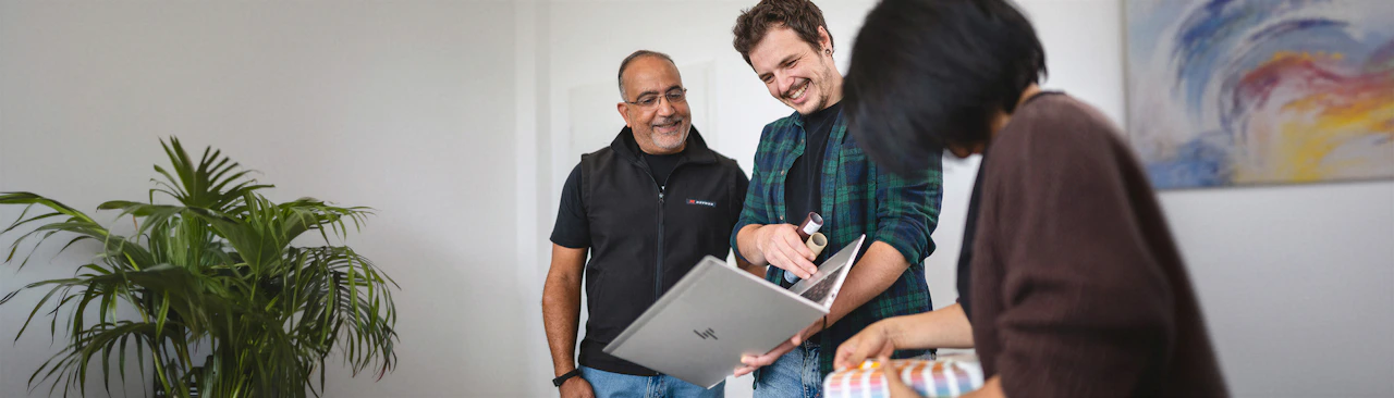 Drei lächelnde Mitarbeiter des Raymon Creative Teams besprechen ein Projekt an einem HP Laptop.
