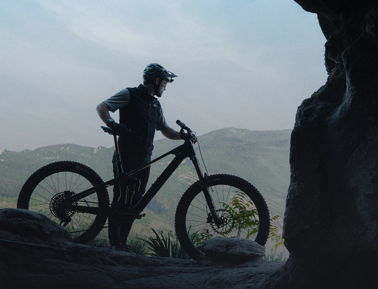 Mountainbiker steht mit seinem Raymon E-MTB an einem felsigen Aussichtspunkt im Gebirge.